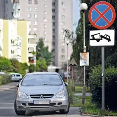 Srebrny samochód na drodze wewnętrznej, obok znaki zakazu zatrzymywania się i holowania. W tle bloki mieszkalne.