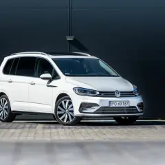 Biały Volkswagen Touran z czarnym dachem i przyciemnianymi szybami, zaparkowany przed ciemną, nowoczesną ścianą.