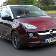 Czerwony Opel Adam z białym dachem jedzie po drodze. Kierowca w czapce siedzi za kierownicą.
