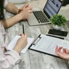 Osoby przy stole omawiają wypowiedzenie umowy ubezpieczenia OC. Na blacie leży laptop, telefon i dokumenty.