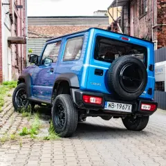 Niebieski Suzuki Jimny z zapasowym kołem na tyle, zaparkowany na brukowanej uliczce między ceglanymi budynkami.