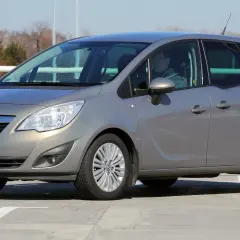 Szary Opel Meriva z kierowcą za kierownicą, zaparkowany na betonowym placu.