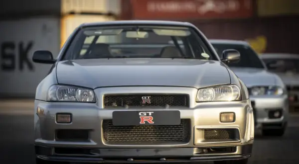 Srebrny Nissan Skyline GT-R R34 z bliska, z innym GT-R w tle. Ikoniczny samochód sportowy.