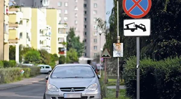 Srebrny samochód na drodze wewnętrznej, obok znaki zakazu zatrzymywania się i holowania. W tle bloki mieszkalne.
