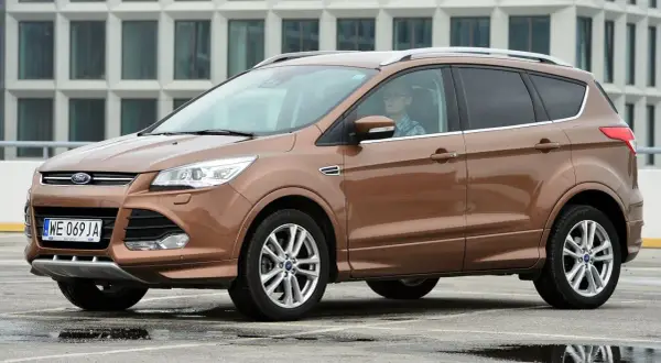 Brązowy Ford Kuga z kierowcą w okularach, zaparkowany na mokrym parkingu przed nowoczesnym budynkiem.