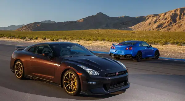 Dwa Nissan GTR R35, jeden brązowy, drugi niebieski, stoją na torze wyścigowym na tle pustynnego krajobrazu.