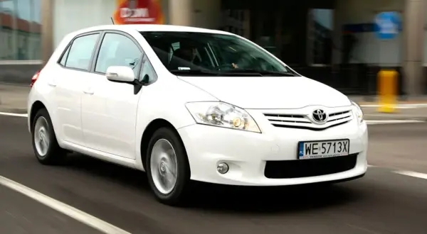 Biała Toyota Auris w ruchu miejskim, z widocznym kierowcą za kierownicą.