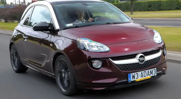 Czerwony Opel Adam z białym dachem jedzie po drodze. Kierowca w czapce siedzi za kierownicą.