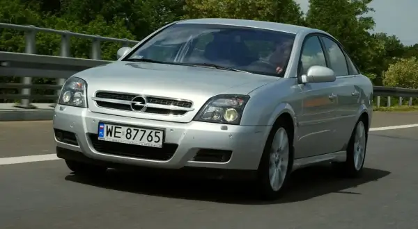 Srebrny Opel Vectra jedzie po drodze, za nim zielone drzewa.