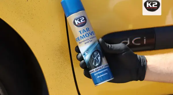Ręka w czarnej rękawiczce trzyma puszkę K2 Tar Remover, która skutecznie usuwa smołę z żółtego lakieru samochodu.