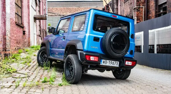 Niebieski Suzuki Jimny z zapasowym kołem na tyle, zaparkowany na brukowanej uliczce między ceglanymi budynkami.