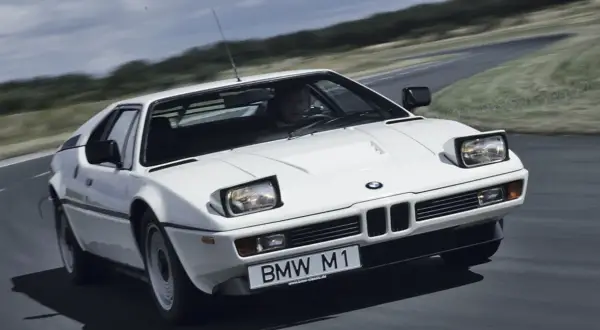 Białe BMW M1 na zakręcie drogi, z widocznym kierowcą i tablicą rejestracyjną.