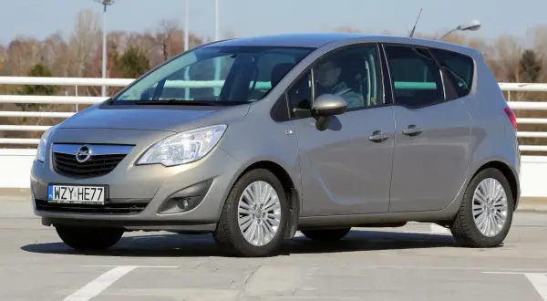 Szary Opel Meriva z kierowcą za kierownicą, zaparkowany na betonowym placu.