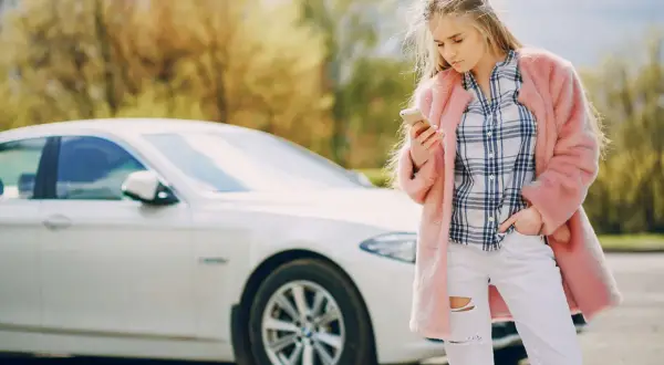 Młoda kobieta w różowym futrze i podartych dżinsach sprawdza telefon obok białego samochodu, być może po **szkodzie parkingowej**.
