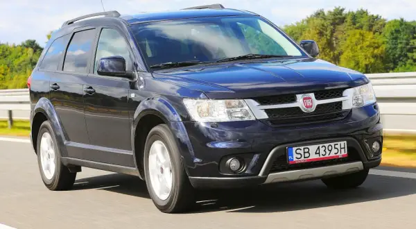 Ciemnoniebieski Fiat Freemont jedzie po drodze, otoczony zielenią drzew i błękitnym niebem.