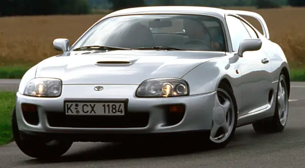 Srebrna Toyota Supra MK4 z charakterystycznym wlotem powietrza na masce, pędząca po asfaltowej drodze wśród pól.
