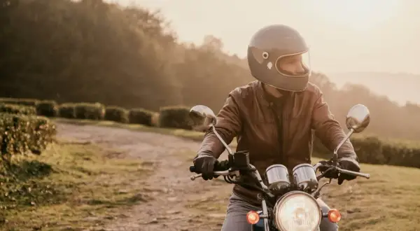 Mężczyzna w kasku i skórzanej kurtce jedzie na motocyklu po polnej drodze. Słońce zachodzi, tworząc ciepłą, złotą poświatę.