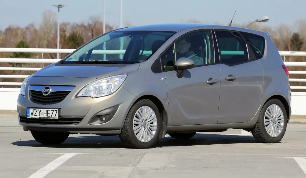 Szary Opel Meriva z kierowcą za kierownicą, zaparkowany na betonowym placu.