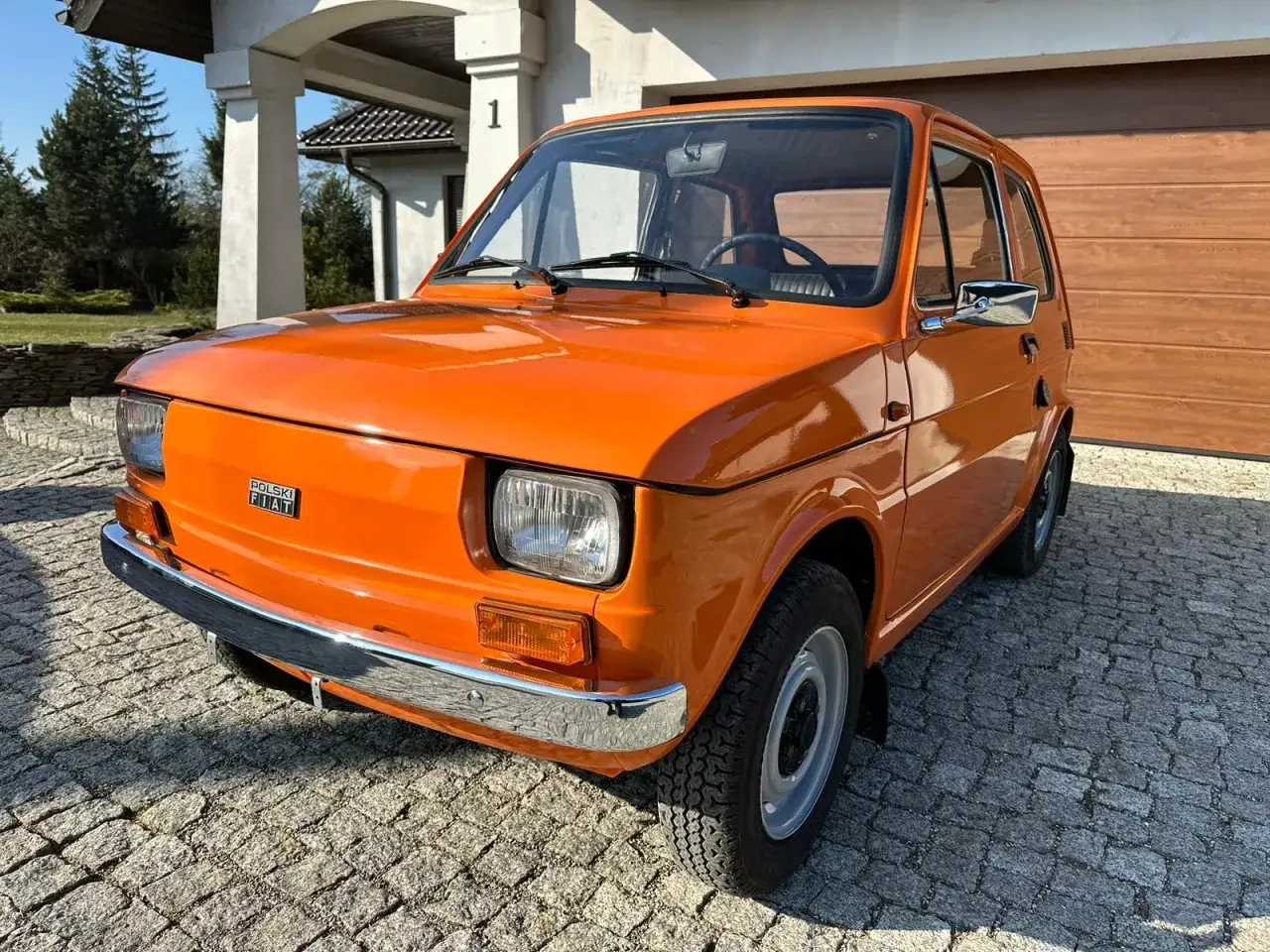 Pomarańczowy fiat 126p lśni w słońcu przed domem. Klasyk na polskich drogach.