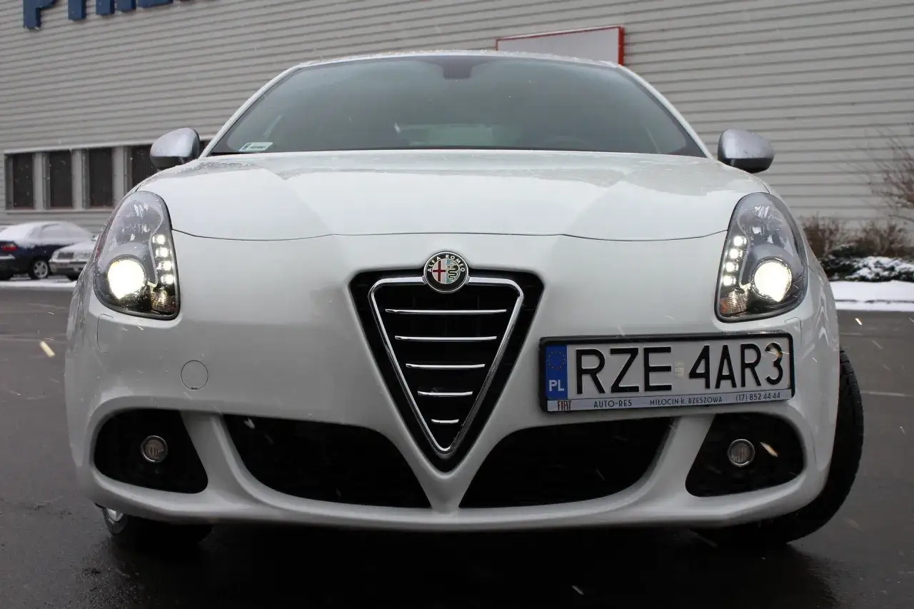 Biała Alfa Romeo Giulietta z włączonymi światłami, na zewnątrz pada śnieg.