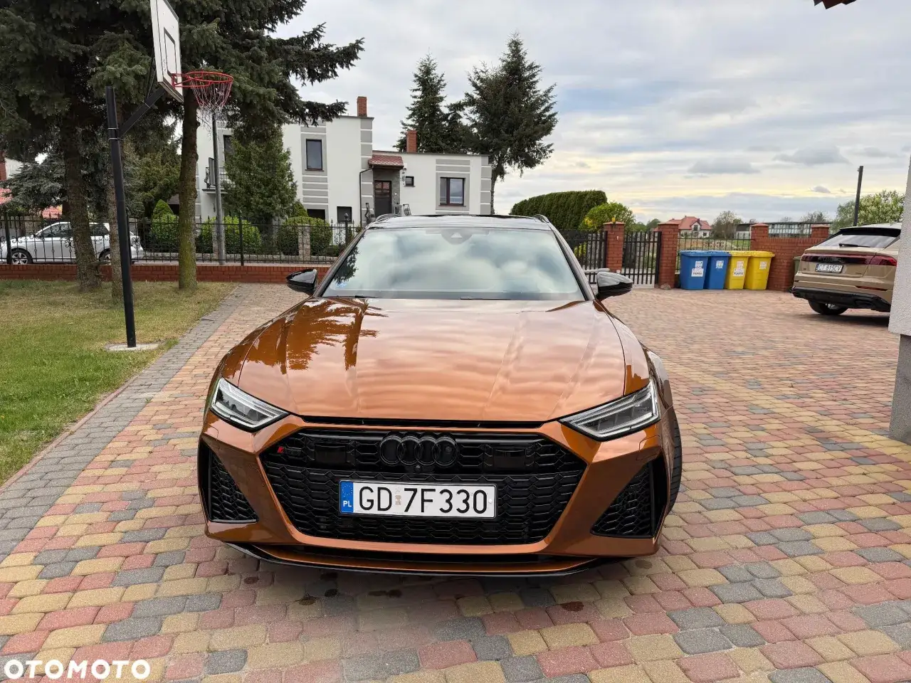 Pomarańczowe Audi RS6 C8 zaparkowane na podjeździe z kolorowej kostki brukowej. W tle dom i kosz do gry w koszykówkę.