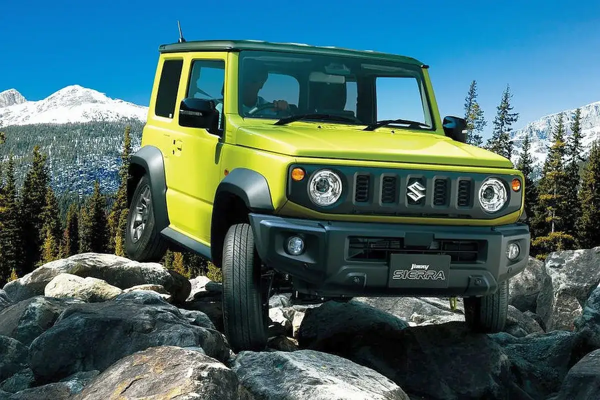 Żółty Suzuki Jimny pokonuje skalisty teren na tle ośnieżonych gór i lasu.