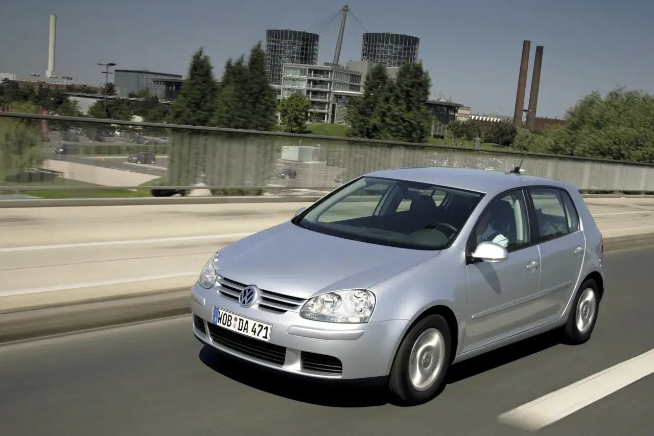 Srebrny Golf 5 jedzie autostradą, w tle nowoczesne budynki i drzewa.