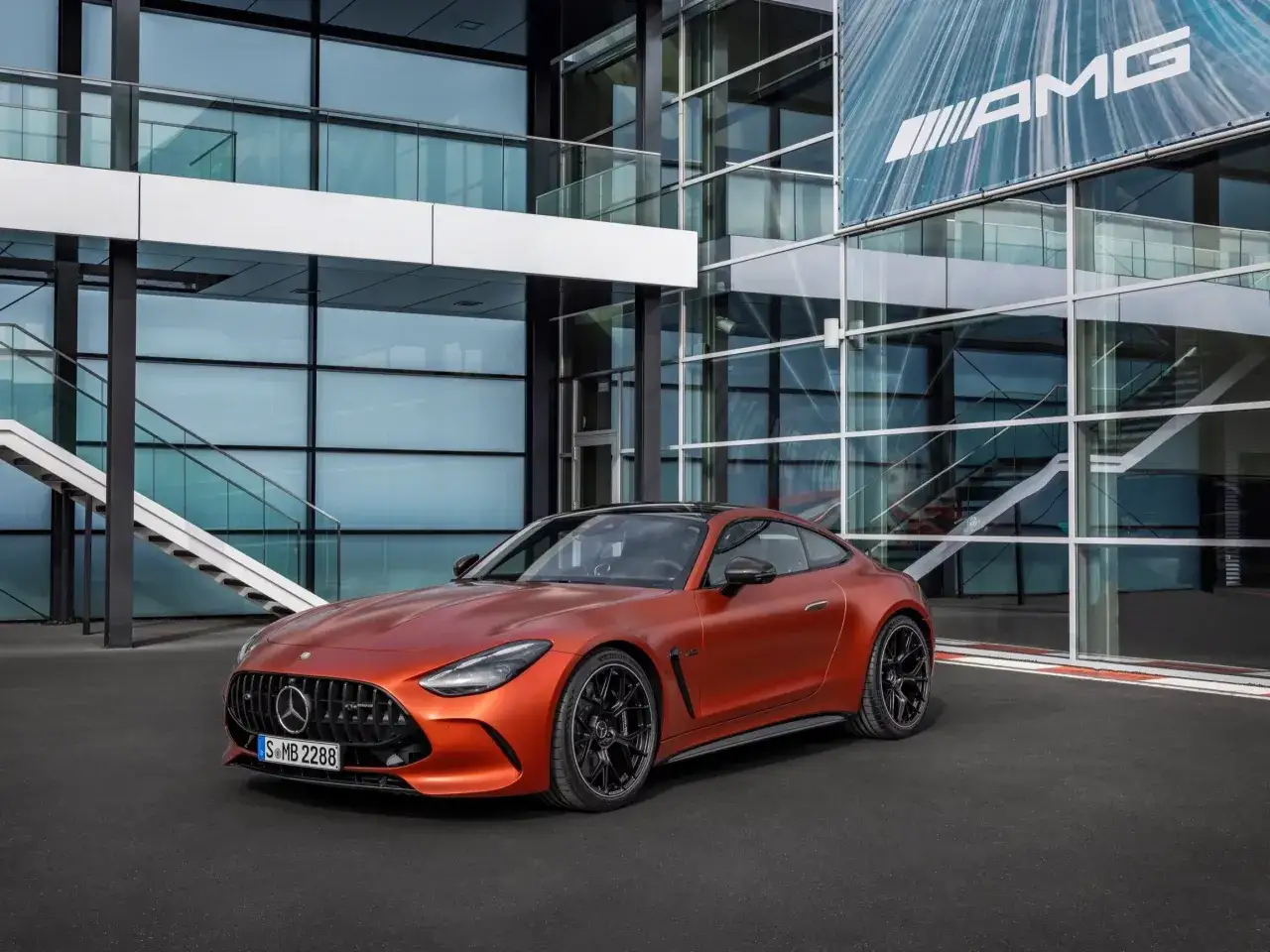 Pomarańczowy Mercedes-AMG GT z czarnymi felgami stoi przed nowoczesnym budynkiem z logo AMG.