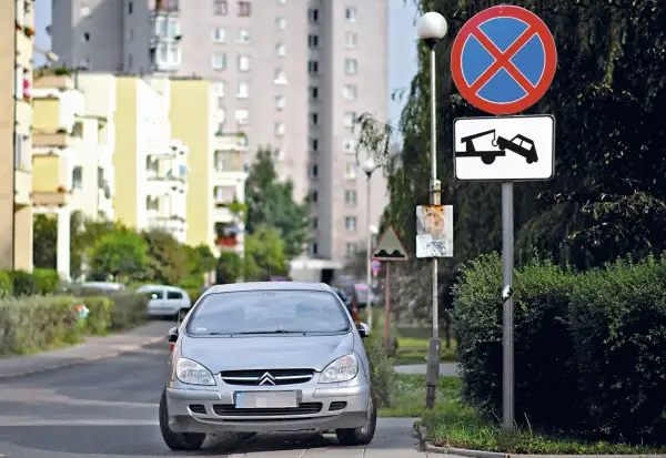 Srebrny samochód na drodze wewnętrznej, obok znaki zakazu zatrzymywania się i holowania. W tle bloki mieszkalne.